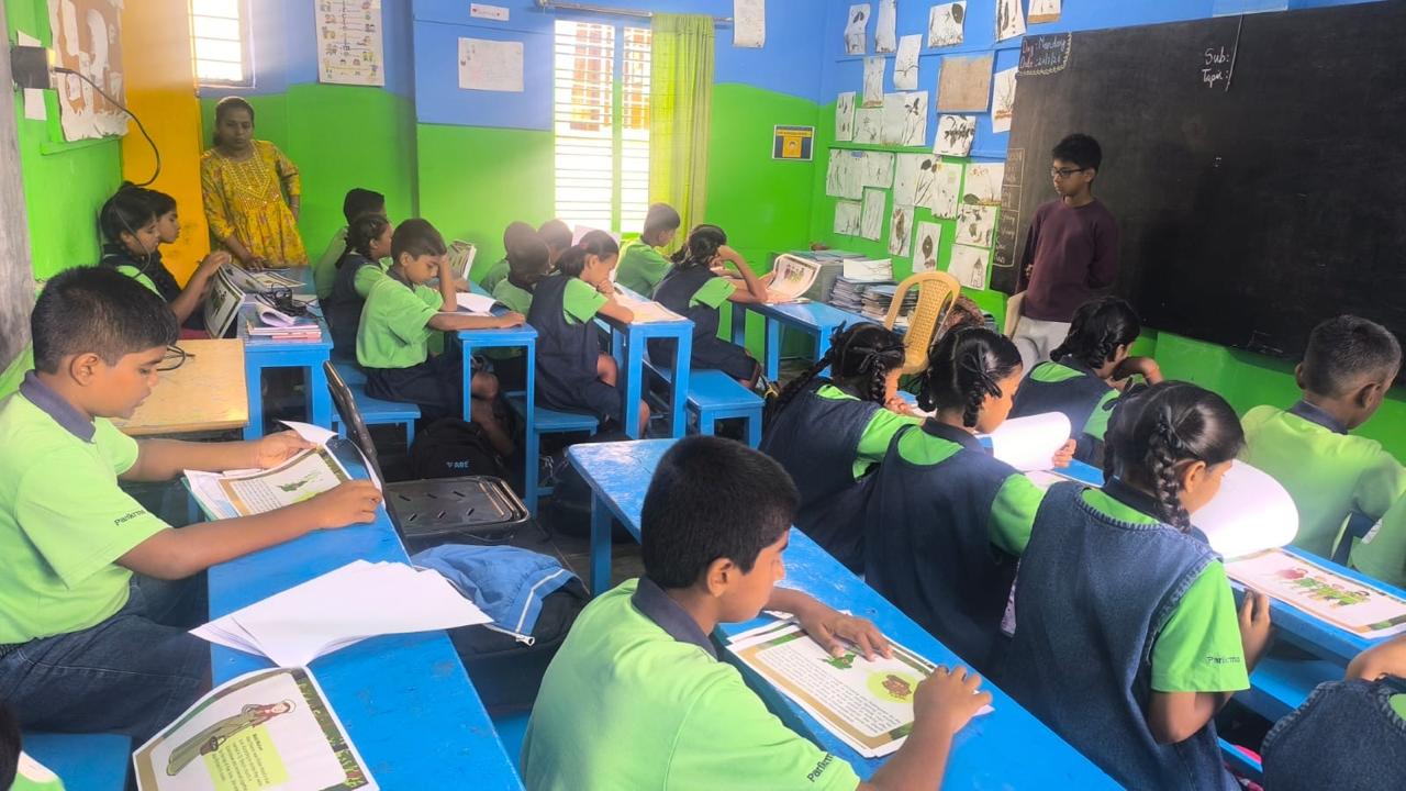 Students reading printed story sheets at their desks while Vanij stands at the blackboard