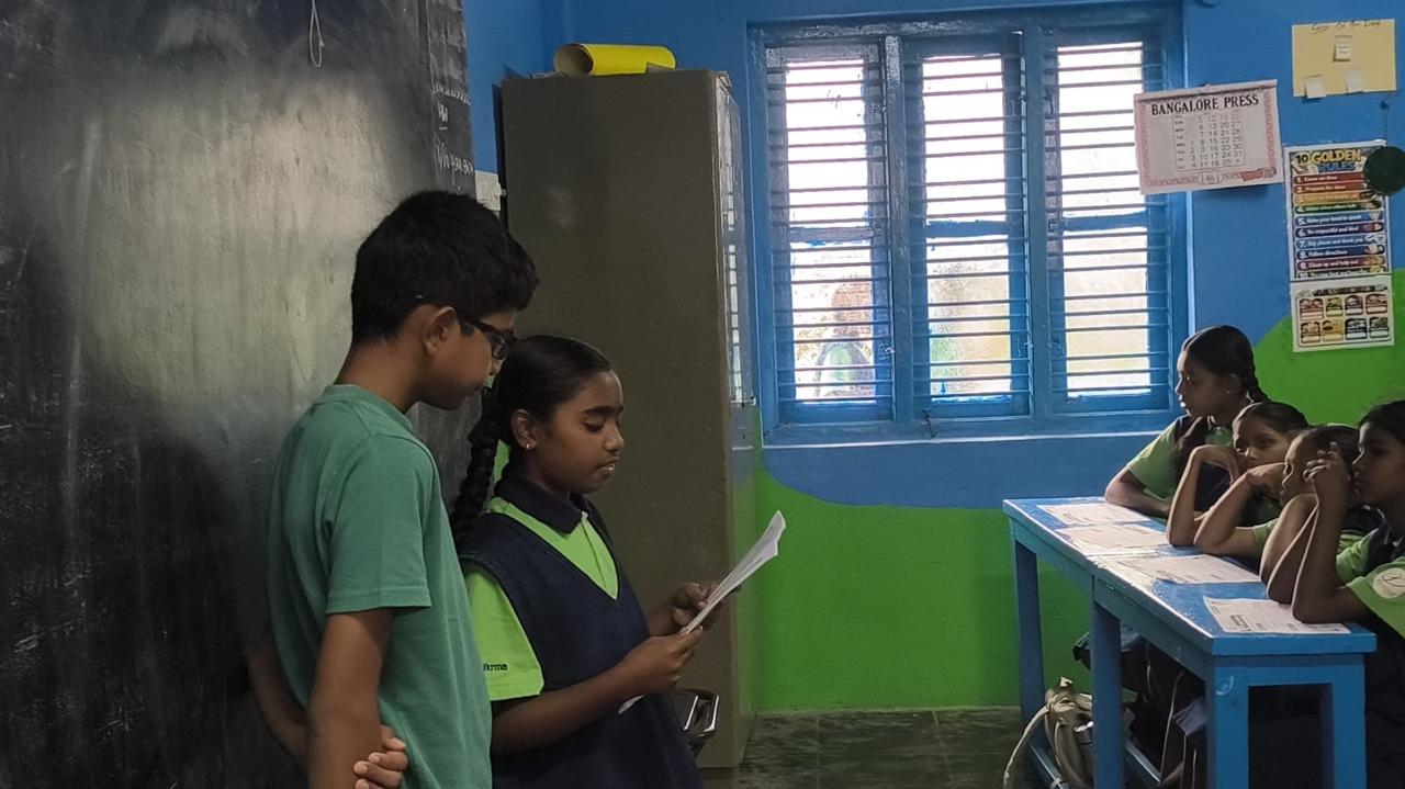 A student reading from a sheet beside Vanij while classmates listen